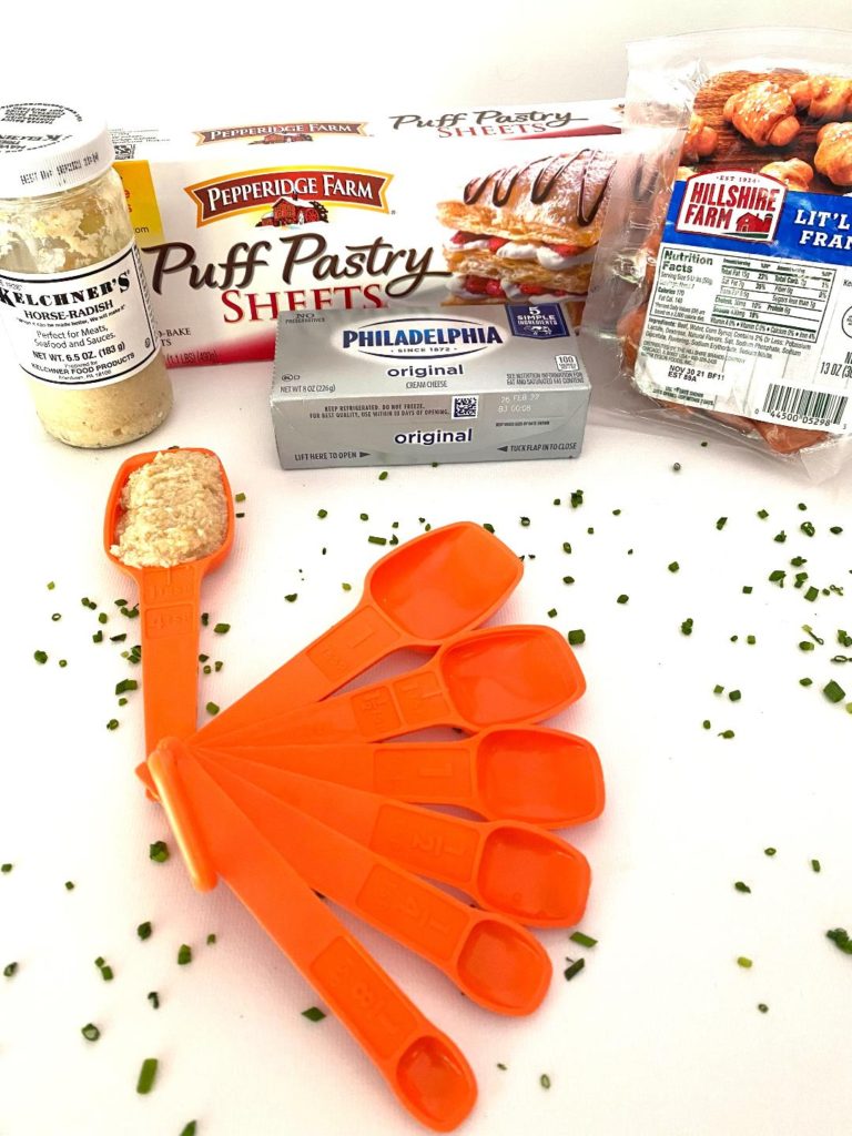Ingredients shown are horse radish in orange vintage tupperware measuring spoons, puff pastry sheets, little franks, cream cheese.